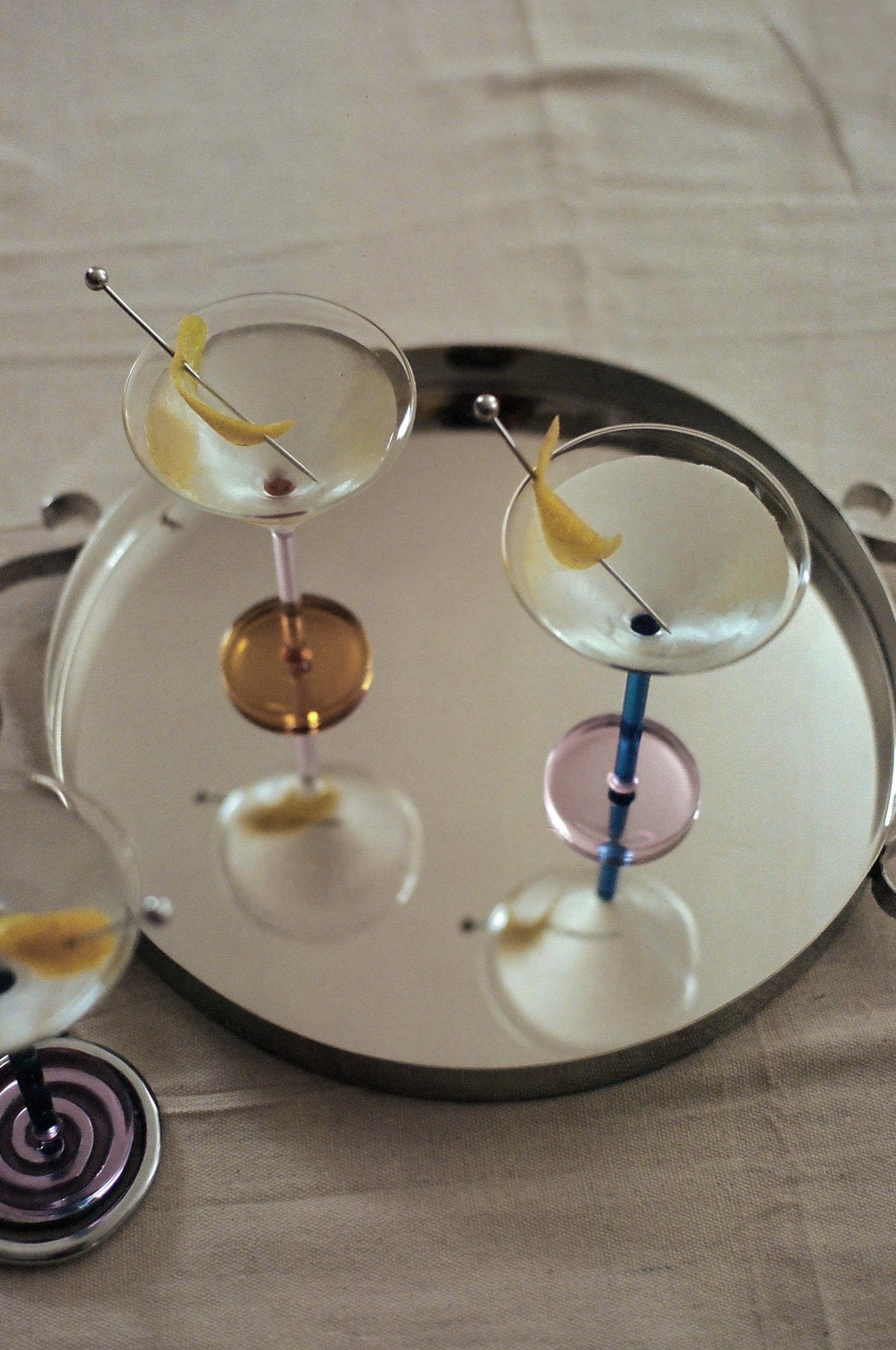 A tray holds three Piano Cocktail Glasses by Sophie Lou Jacobsen, each with a lemon twist; the glasses feature colorful stems, including yellow, pink, and purple, reflected on the tray's surface.