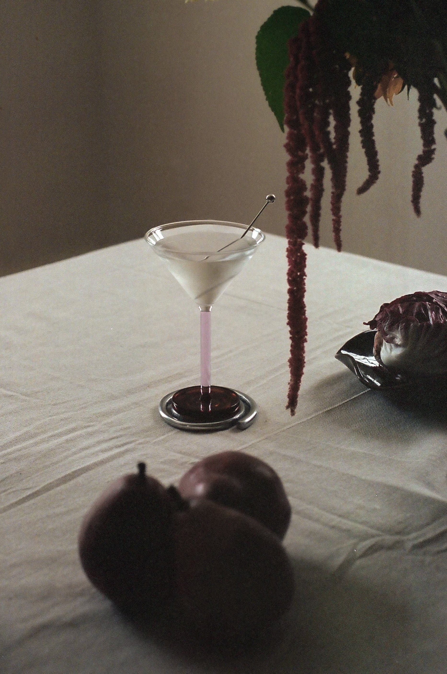 A still life featuring a Piano cocktail glass with a pink stem on a table, accompanied by three purple plums and draped amaranth flowers on the side. The table is covered with Sophie Lou Jacobsen.