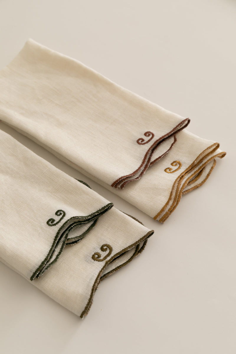 Three folded Sophie Lou Jacobsen Twist Napkins on a light background, each with a different colored embroidered design at the corner: maroon, ochre, and green, perfect for a fine dining table setting.