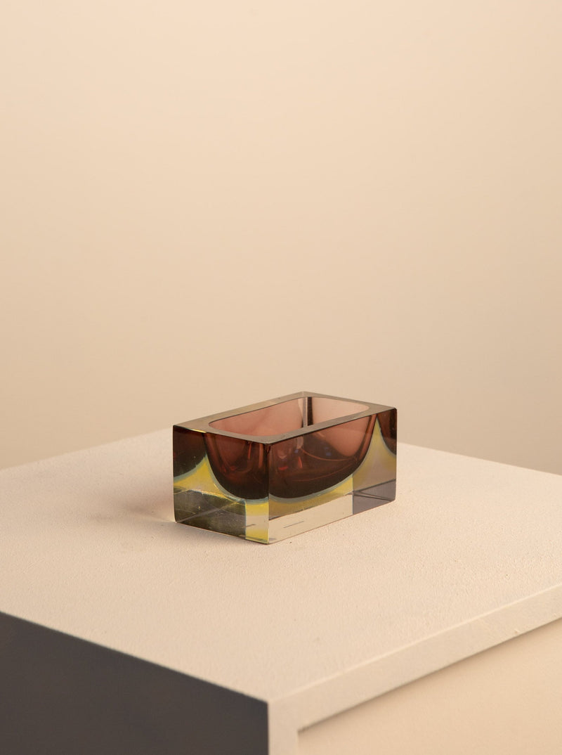 A clear Mauve Murano Vide Poche By Flavio Poli containing a pair of sunglasses with dark lenses placed against a beige background. The glasses appear to float within the block. Brand: Treaptyque