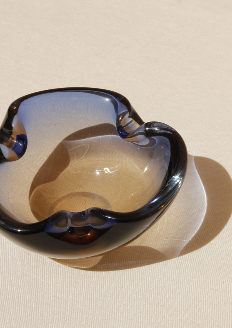 The Two-Colour Murano Bowl from Boga Avante Shop features a wavy, organic shape in a gradient of blue to clear tones. It casts a soft shadow on any beige surface, exemplifying contemporary design as both functional art and collectible furniture.