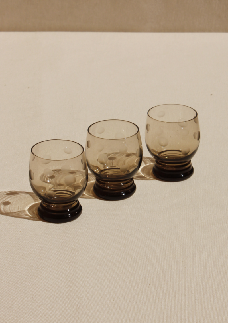 Three empty, vintage Boga Avante Shop polka dot glasses with thick bases, aligned in a row on a beige surface, casting soft shadows.