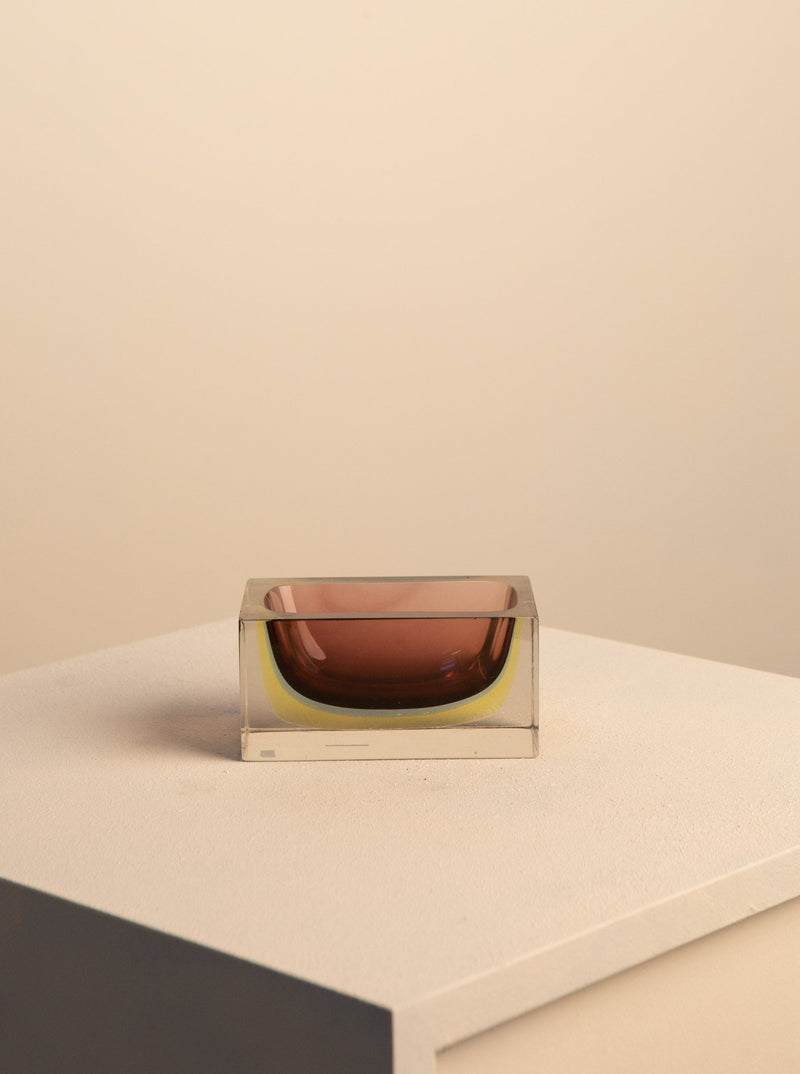 A minimalist image featuring a dark amber-colored, oval-shaped Mauve Murano Vide Poche by Flavio Poli centered within a transparent square block, placed on a light beige surface against a matching backdrop. (Brand Name: Treaptyque)