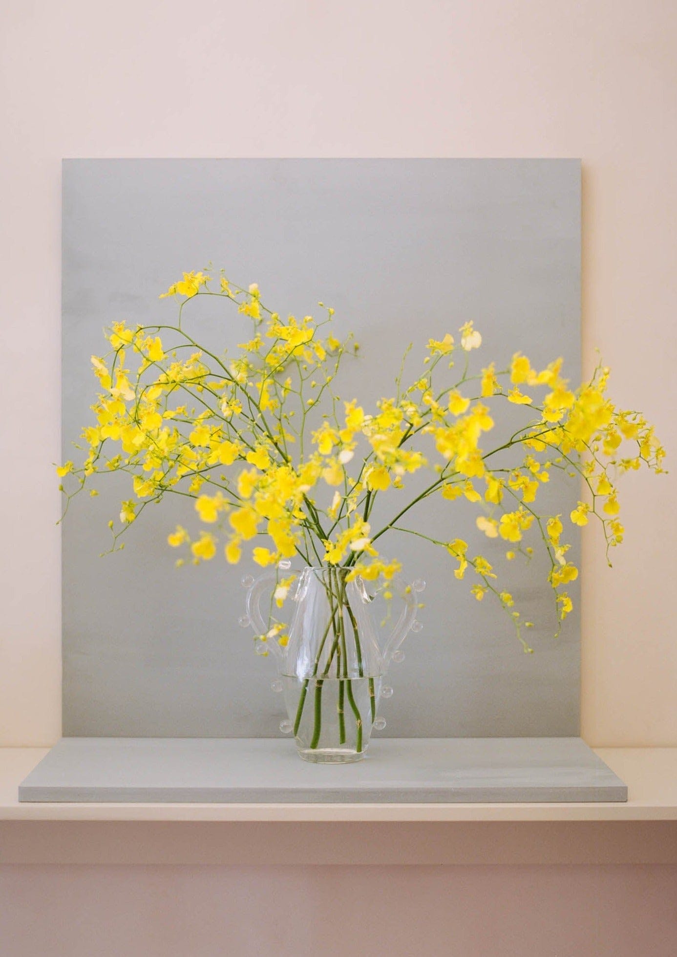 A bright bouquet of yellow flowers arranged in a La Vitalité Vase by Sophie Lou Jacobsen, set against a gray canvas on a light beige shelf.