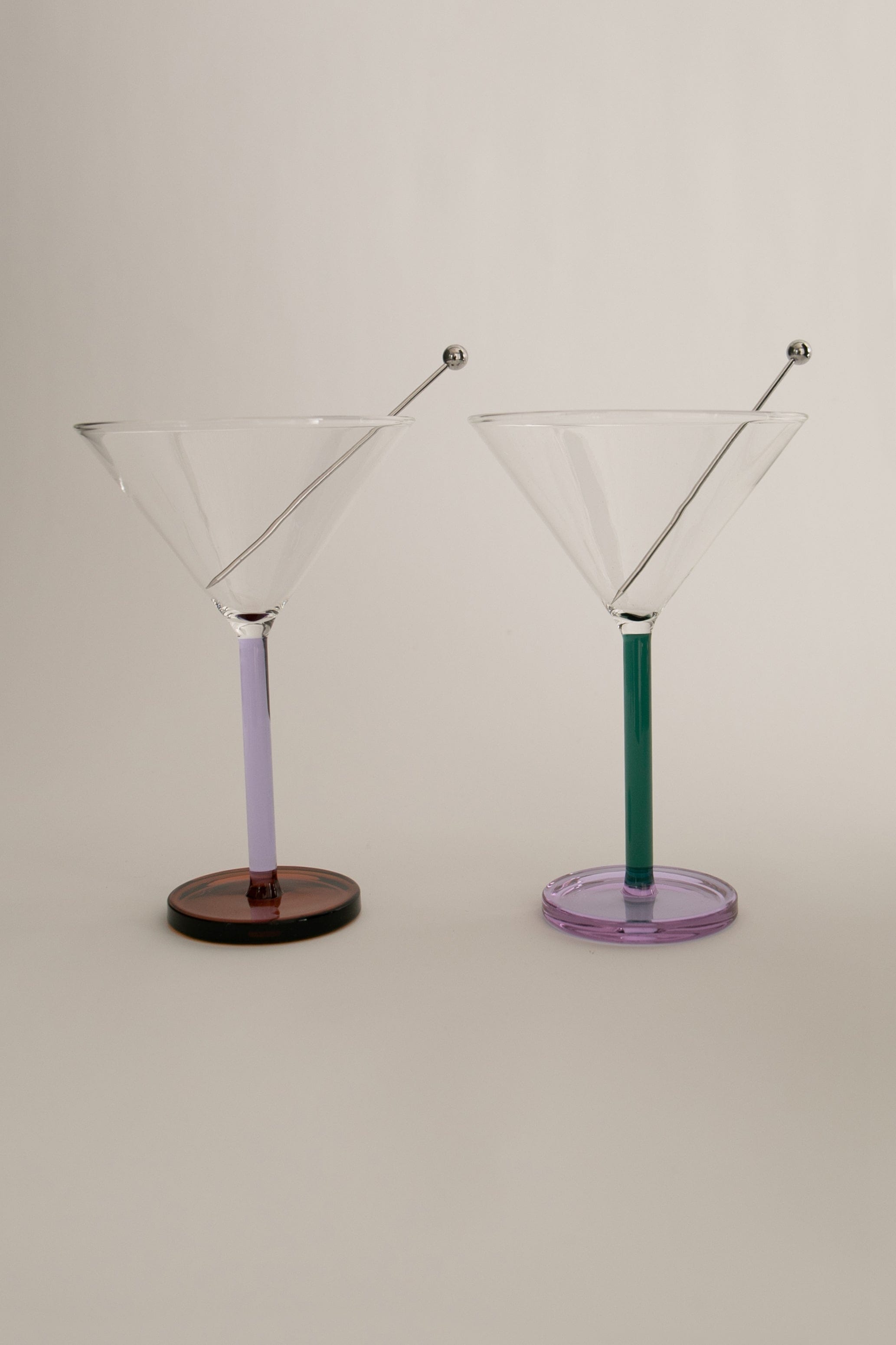 Two Piano Cocktail Glasses with colorful stems, one purple and one green, standing against a neutral background. Each glass features a small, round embellishment at the tip of the stem.
