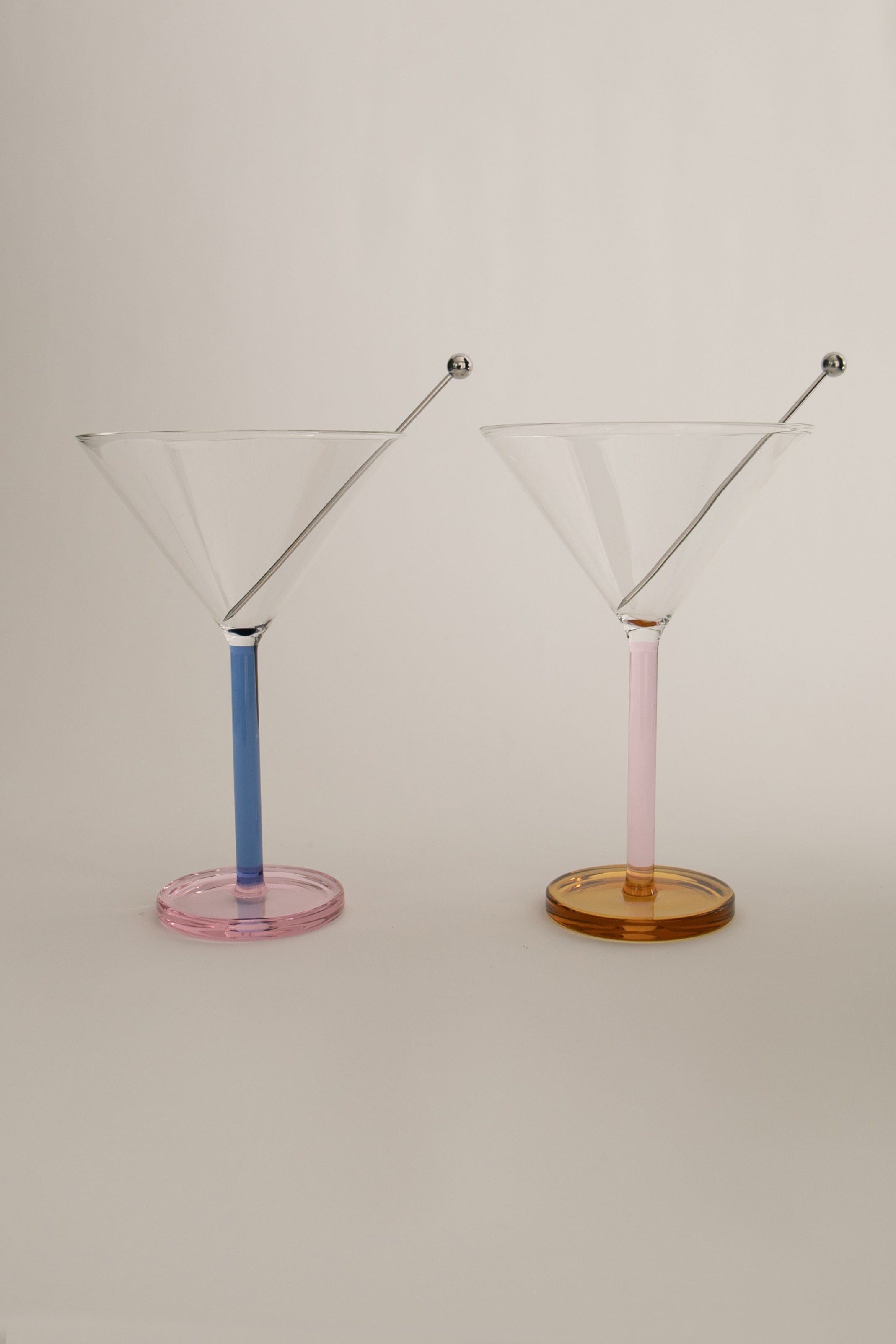 Two Piano cocktail glasses with colorful stems, one pink and one blue, against a light neutral background. The glasses are empty and have stainless steel garnish skewers as decorations on their rims.