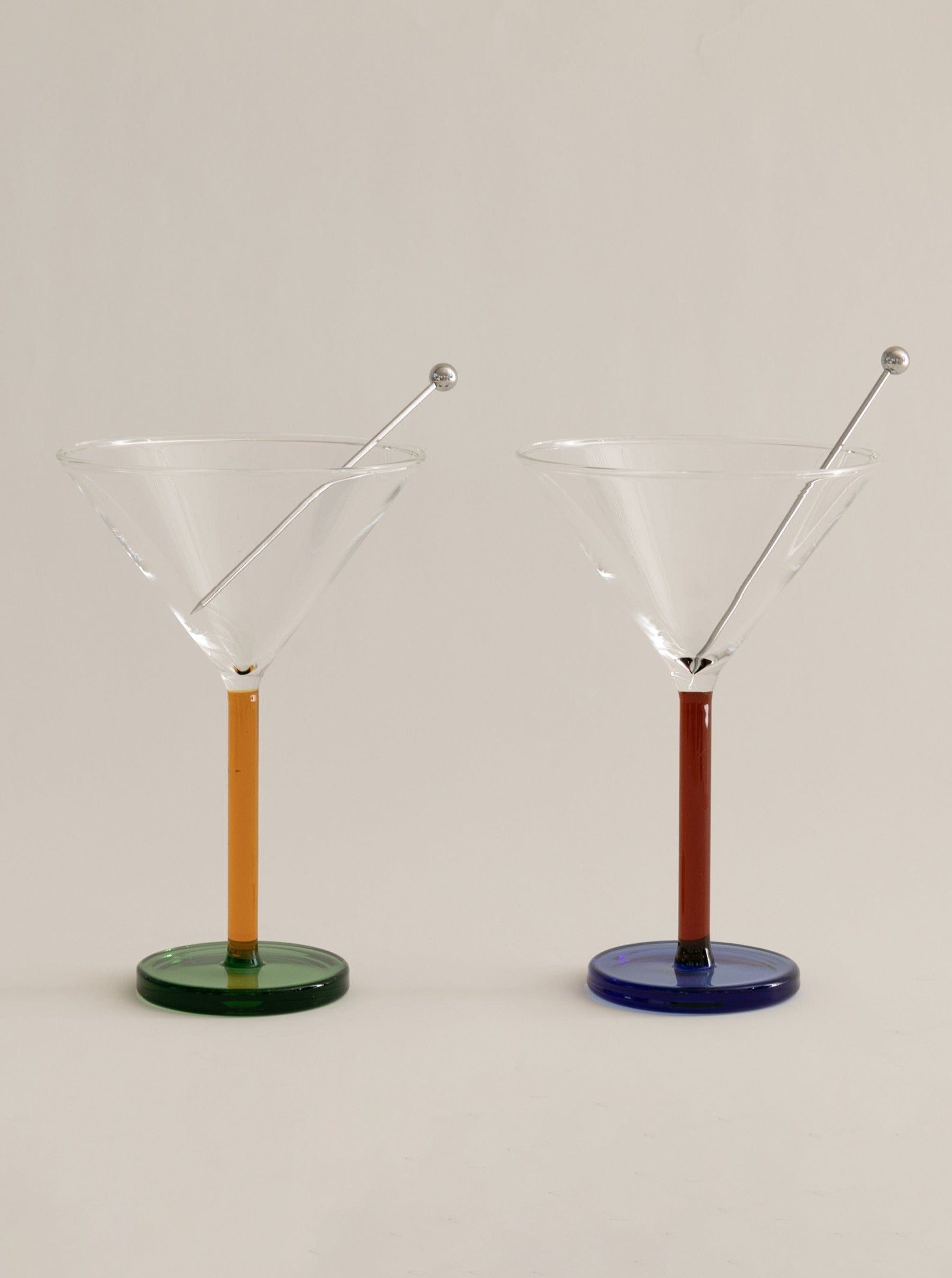 Two elegant Sophie Lou Jacobsen Piano Cocktail Glasses with clear bowls, green and blue bases, and decorative stainless steel garnish skewers on a neutral background.