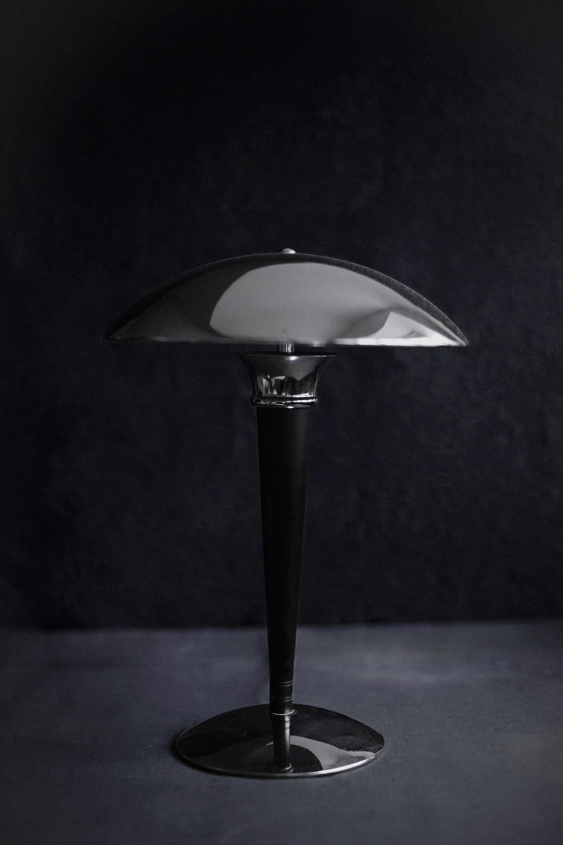 An elegant black Out For Lunch Bauhaus Table Lamp with a sleek, curved lampshade following Bauhaus principles, illuminated from underneath, set against a dark textured background.