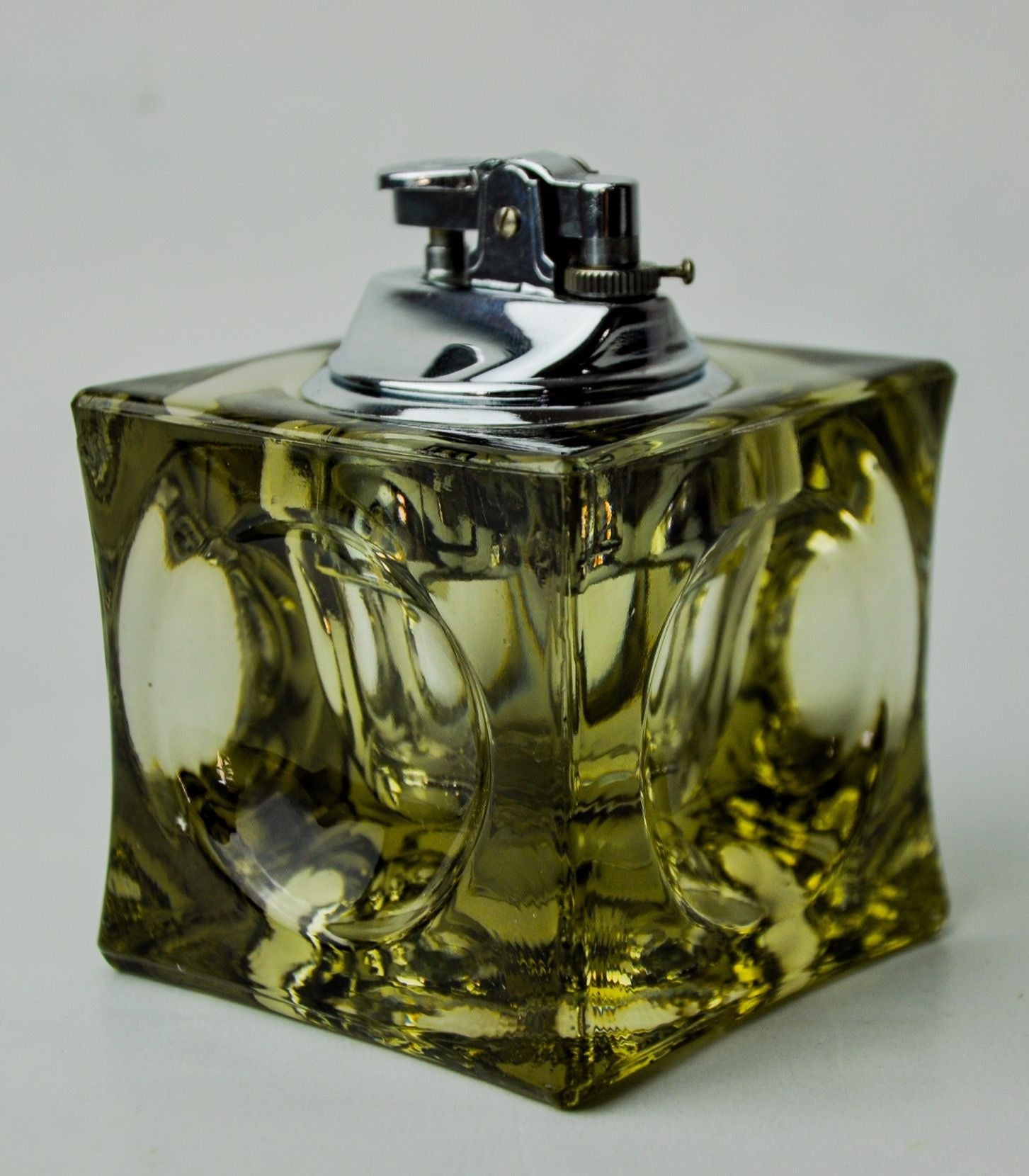 A square Murano glass Ice cube lighter by Antonio Imperatore, 1970, with a silver spray nozzle, filled with yellow liquid, against a light gray background. The glass has a unique embossed design.