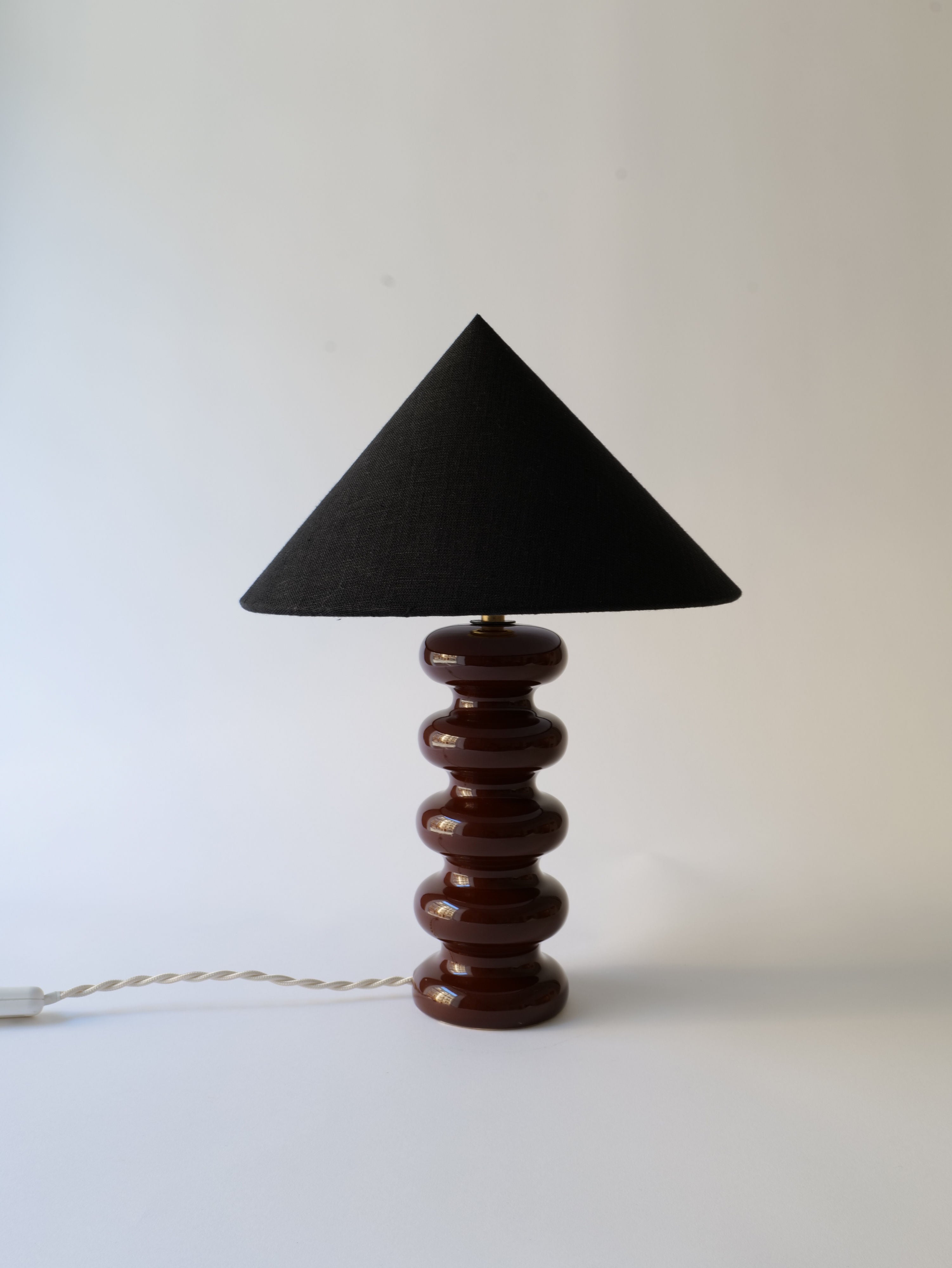A modern Sculptural Ceramic Lamp with a triangular black shade and a base composed of stacked circular Doulton England ceramic elements, on a white background by Collection apart.