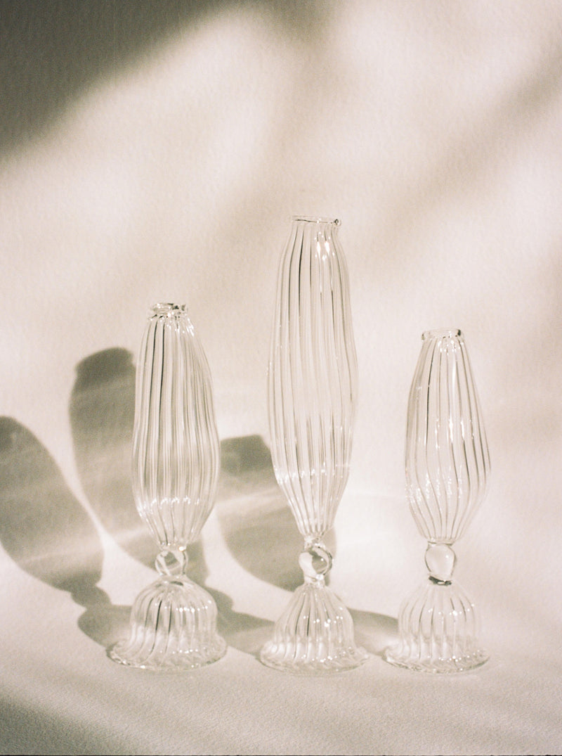 Four elegant mouth-blown glass Three Sisters Vases by Justine Menard with vertical ribbed patterns cast soft, intricate shadows on a draped cloth surface, illuminated by natural light.