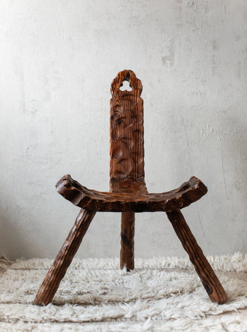A rustic brown Out For Lunch Brutalist Chair with intricate carvings stands on a shaggy white rug against a textured grey wall. The chair features a distinctively shaped backrest and sturdy legs.
