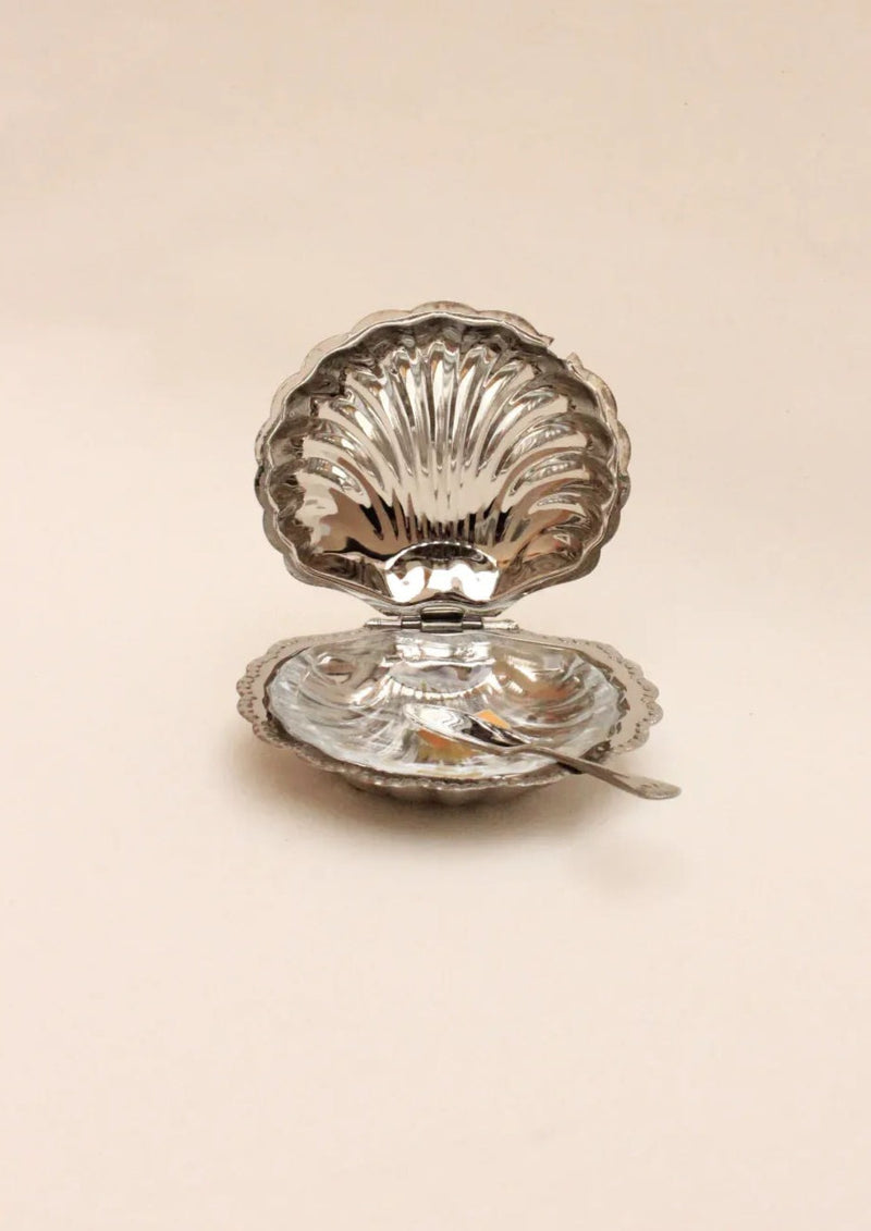 The Boga Avante Shop Shell Shaped Butter Dish, featuring a shiny silver clam design with a sculptural, hinged lid, is open to reveal its minimalist beauty. A small silver spoon elegantly rests inside the handcrafted lower half against a plain neutral background.