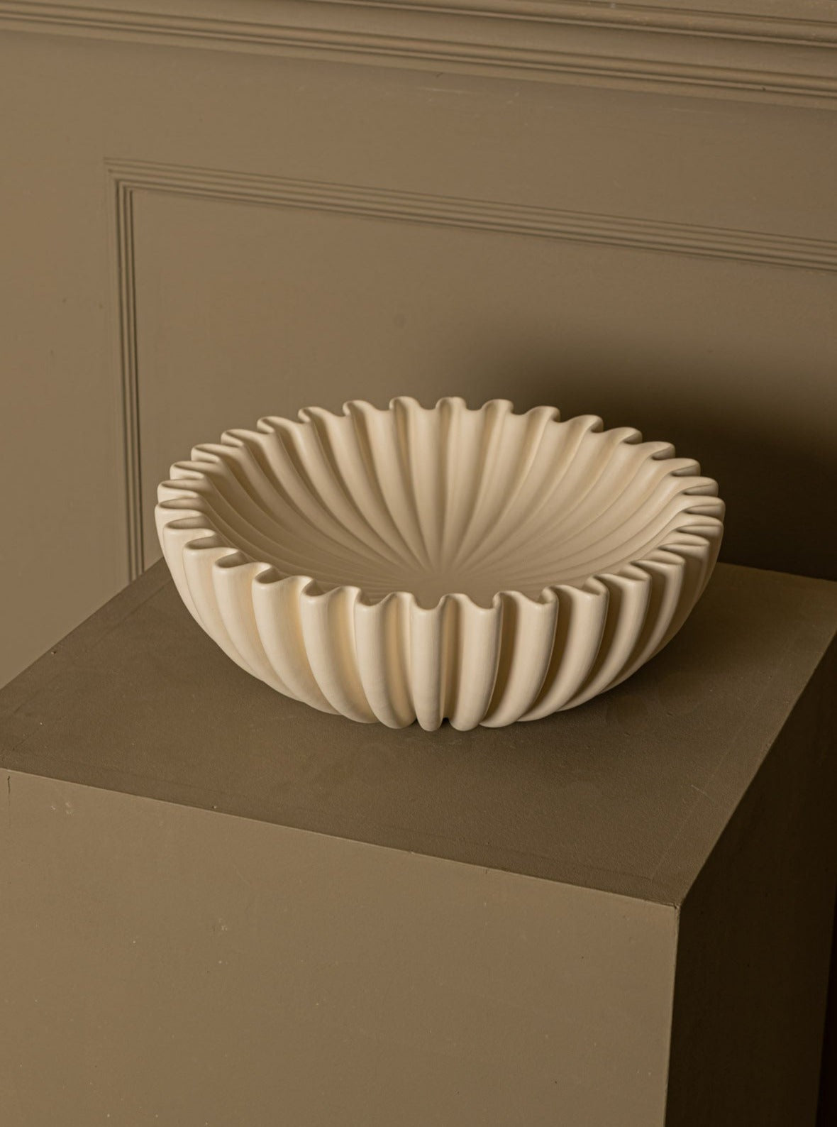 A decorative, fluted Simone & Marcel Lotuso Ecru Ceramic art bowl, placed on a square pedestal against a textured beige wall. The bowl features a beautifully detailed, radial pattern.