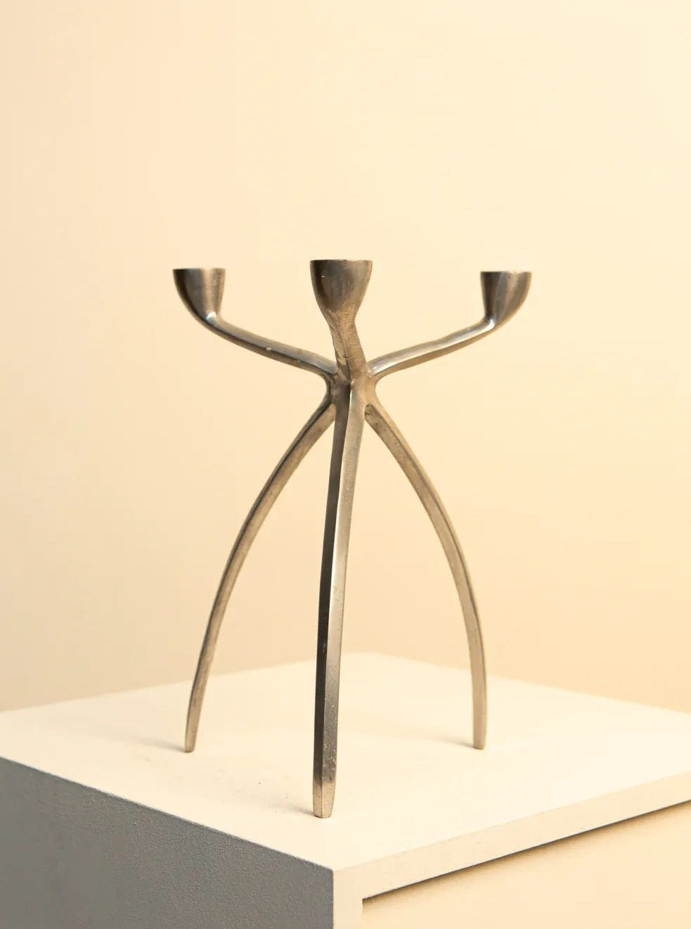 Abstract metal sculpture resembling a human figure with arms stretched upwards, holding a Treaptyque 70's Metal "Tripod" Candleholder, displayed on a white pedestal against a soft beige background.