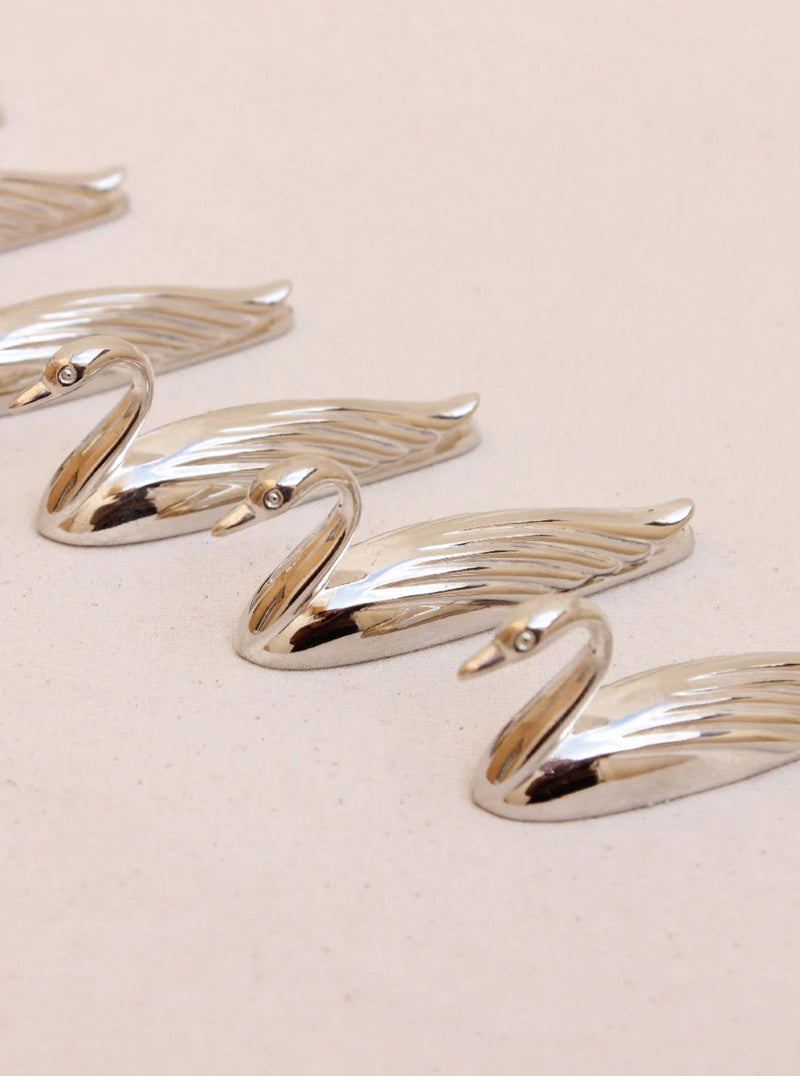 The Set of Swan Cutlery Holders by Boga Avante Shop is elegantly displayed on a light beige surface. With detailed feather engravings and elongated necks, these pieces bring luxury and sophistication to any setting.
