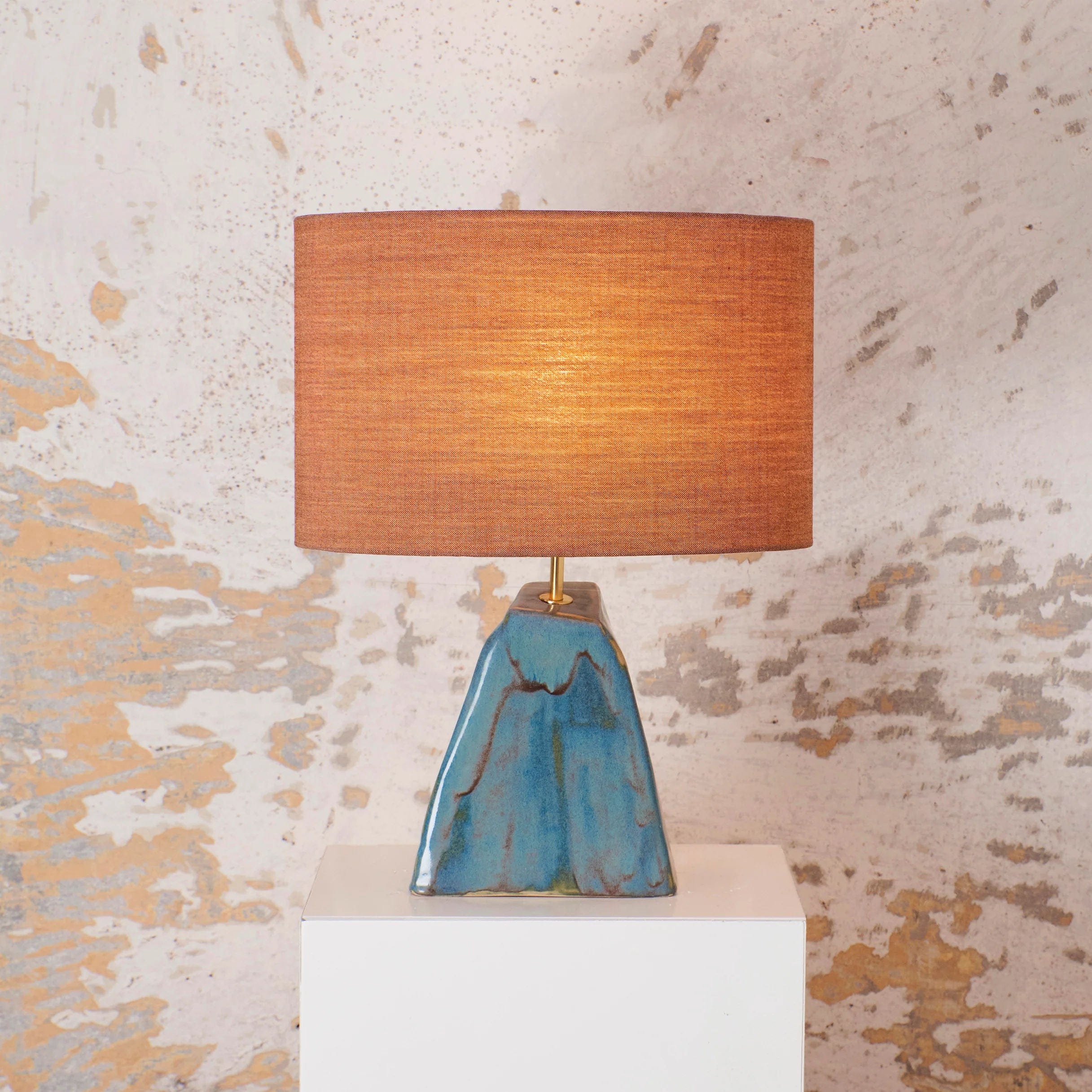 A stylish Triangular Ceramic Lamp by Project 213A with a blue and teal ceramic base and a cylindrical orange shade, illuminated and placed on a white pedestal against a textured beige wall, showcasing artisanal craftsmanship.