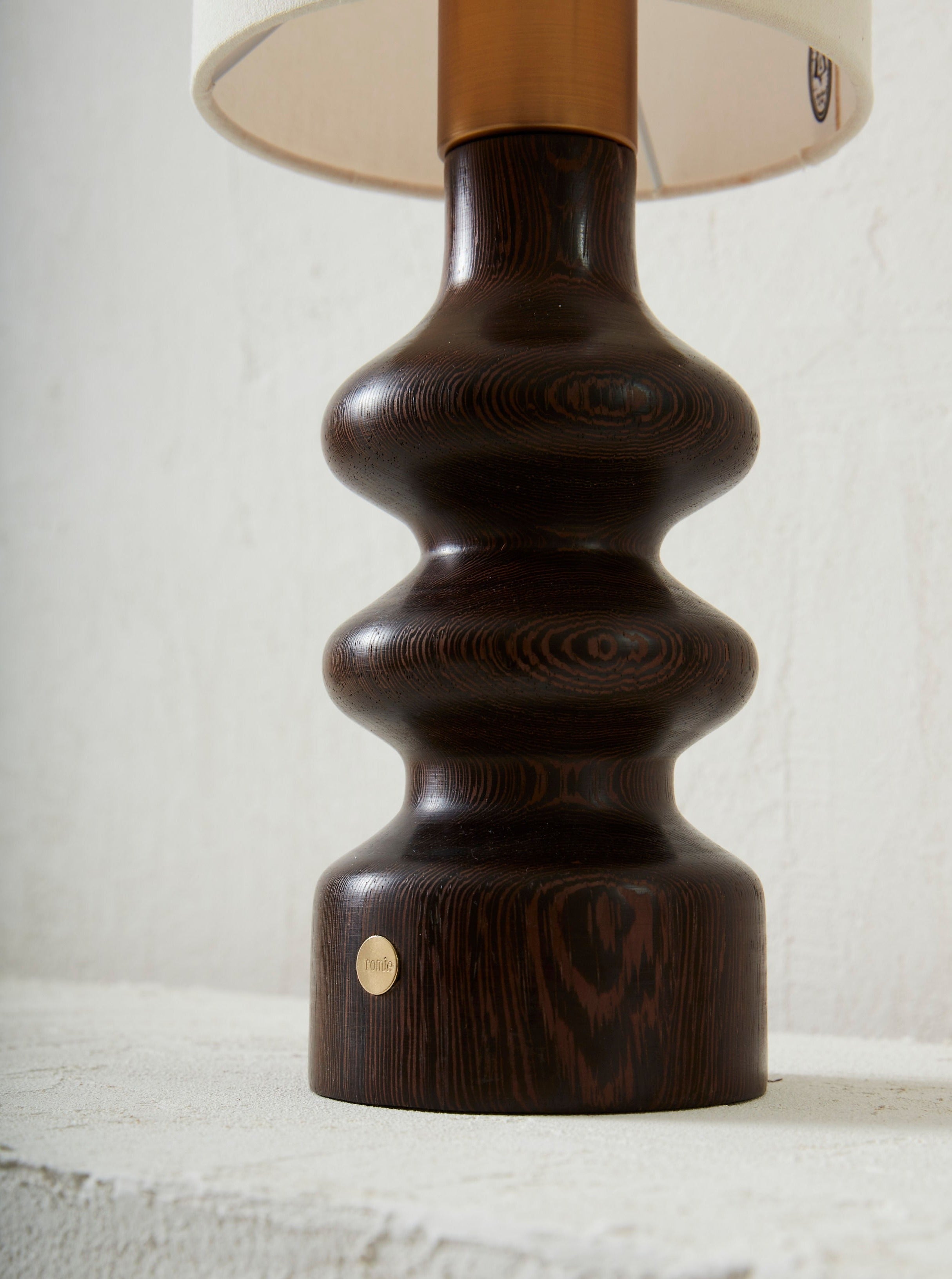 Close-up of a dark wenge grain textured Romie Objetti Alma Wengé Lamp base with a smooth, curvy design, featuring a small, round, brass-colored brand plaque on its lower portion. A cream lampshade is.