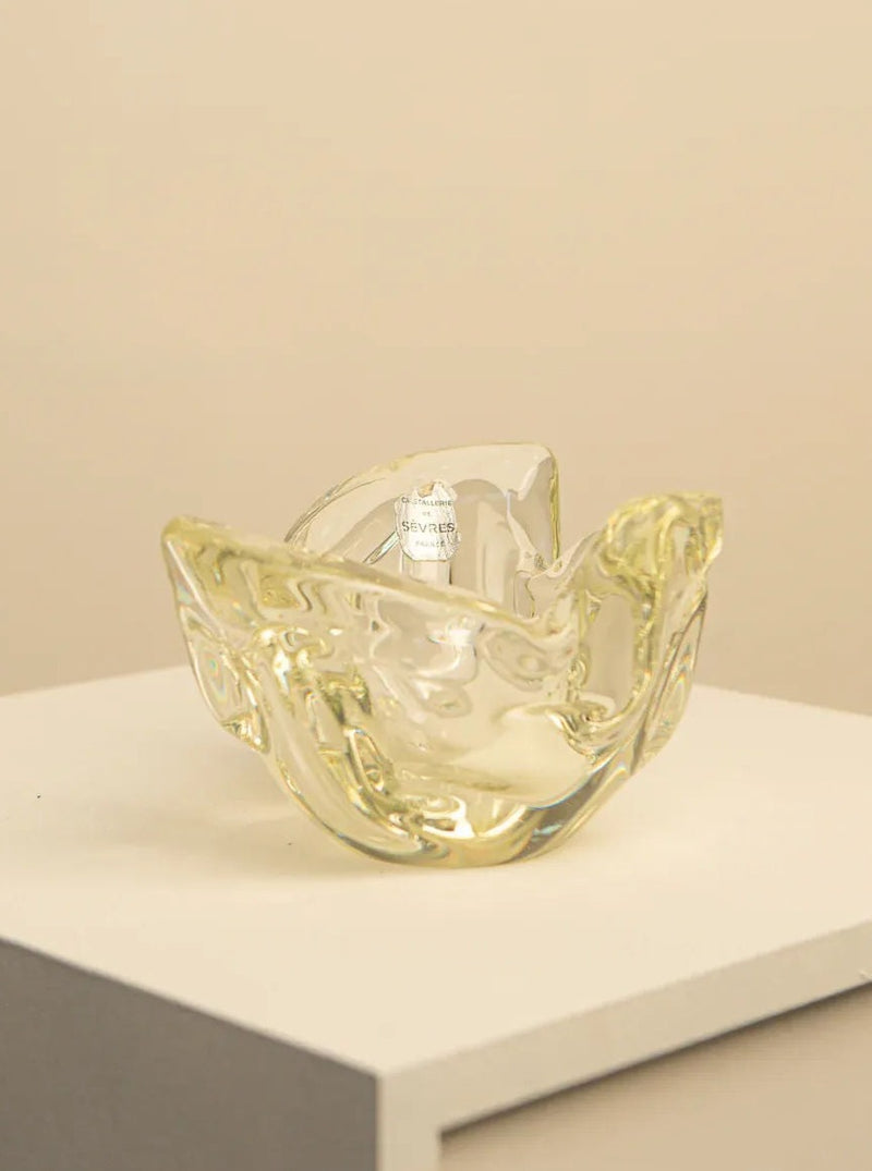 The Lemon Yellow Crystal Bowl by Sèvres 60's, a stunning creation by Treaptyque, rests elegantly on a white pedestal against a beige background. This modern decorative piece boasts a unique, abstract folded design with smooth, undulating surfaces and reflective highlights.