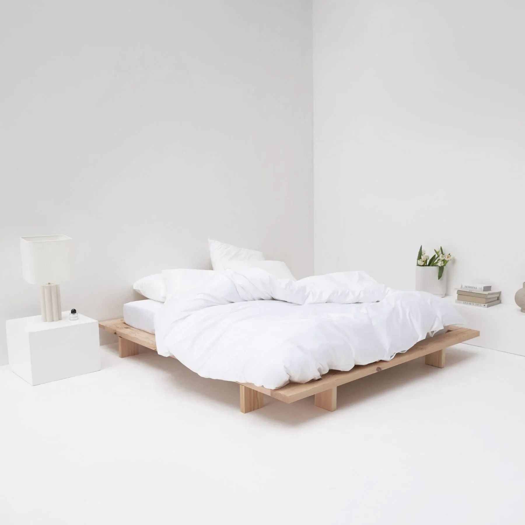 A minimalist bedroom with a low wooden bed, MOST white cotton sateen bed set, a white floor and walls, featuring a small bedside table, lamp, and a few books and plants.