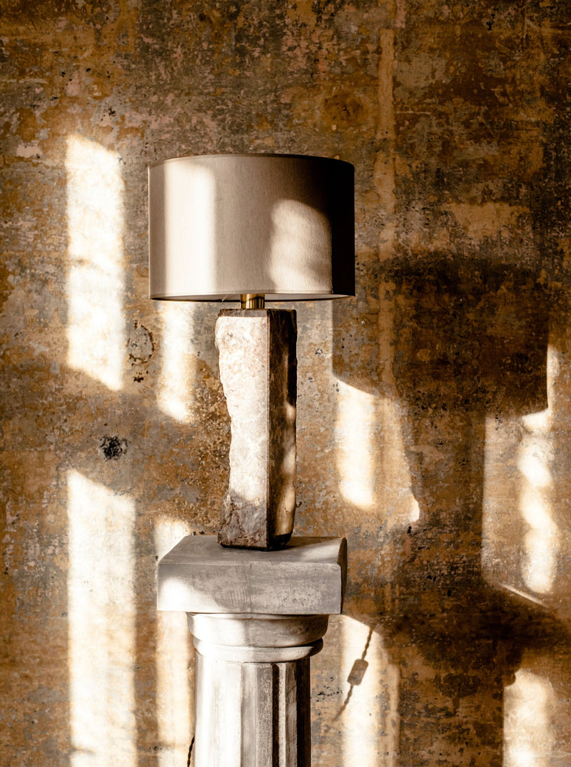 A Pompeii Lamp 1 with a beige shade balances on a rustic, textured Brajak Vitberg marble and wooden pedestal against a concrete wall with sunlit patterns.