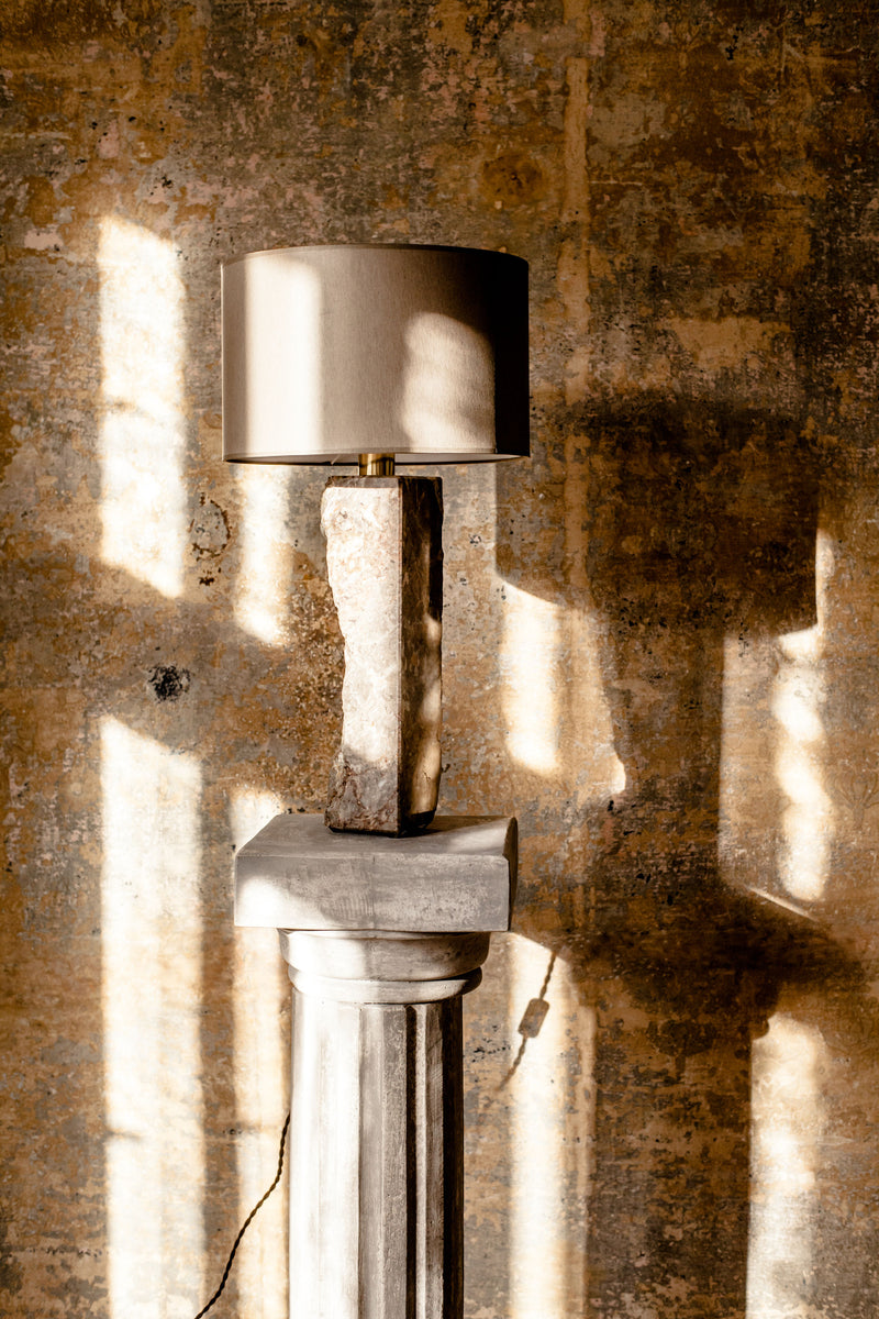 A Pompeii Lamp 1 by Brajak Vitberg with a cylindrical shade sits atop a classical stone column made from Podpeški Slovenian marble, bathed in dappled sunlight casting striped shadows on a textured wall.