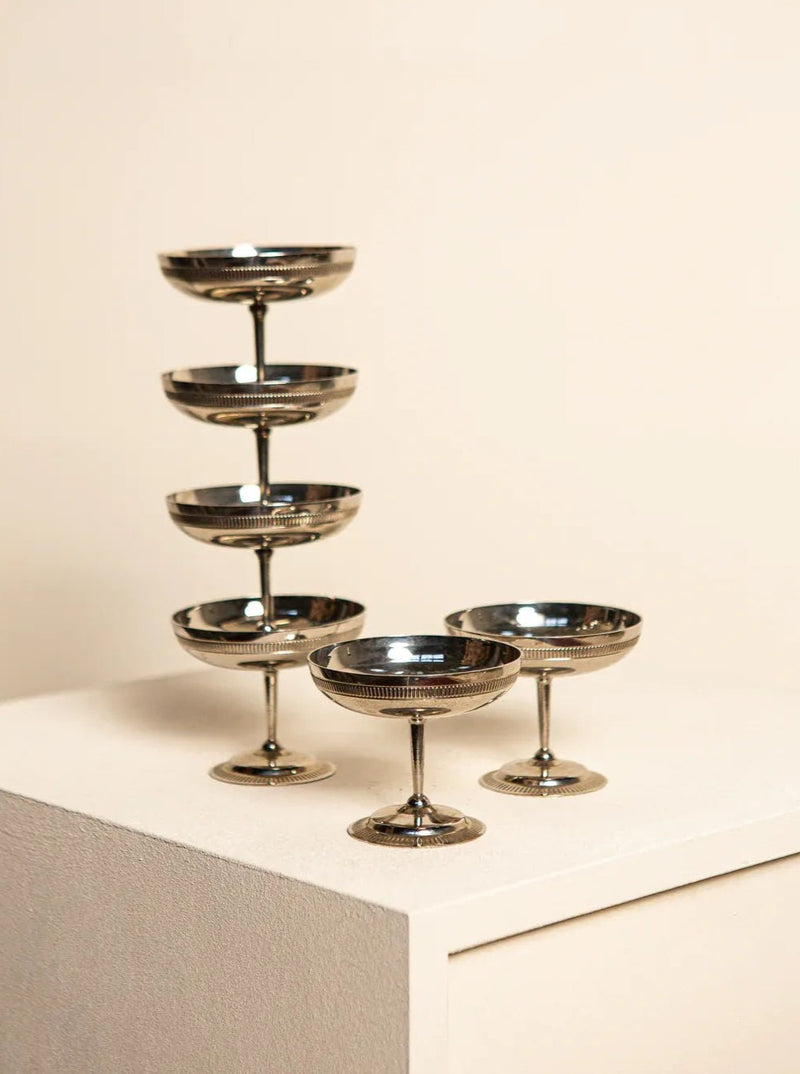 A set of elegant, Treaptyque metallic champagne coupes arranged aesthetically on a cream-colored surface, exuding retro elegance with a tower of stacked coupes in the background.