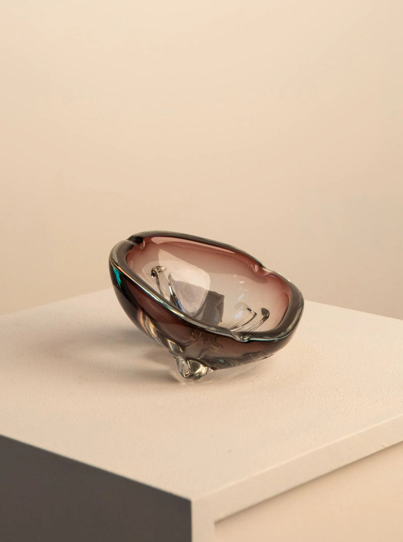 An elegant Treaptyque 70's Mauve Murano Glass Ashtray with a dark amber hue rests on a white pedestal against a light beige background, showcasing its smooth, reflective surface and unique design.