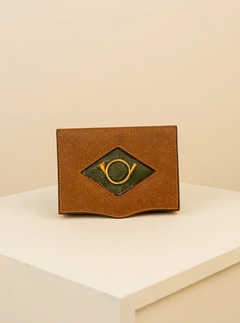 A small, brown Treaptyque leather wallet with a distinctive green and gold eye-shaped emblem, displayed on a white pedestal against a neutral background.