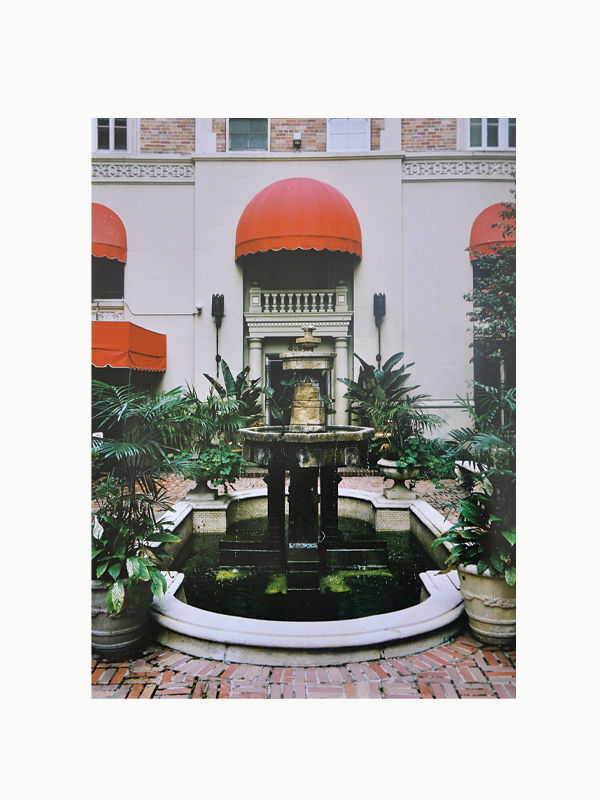 A serene courtyard featuring an elegant Maison Plage Art Deco style fountain centered between lush green plants, flanked by two red awnings atop stone building archways.