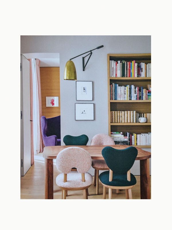 A cozy dining area featuring a wooden table surrounded by uniquely designed chairs with pastel-colored cushions. In the background is a built-in bookshelf filled with books, including "The Orange Set of 3 Interior Design and Art Books" by Maison Plage for creative inspiration, and a wall-mounted lamp next to two framed pictures, with a glimpse of a purple armchair.