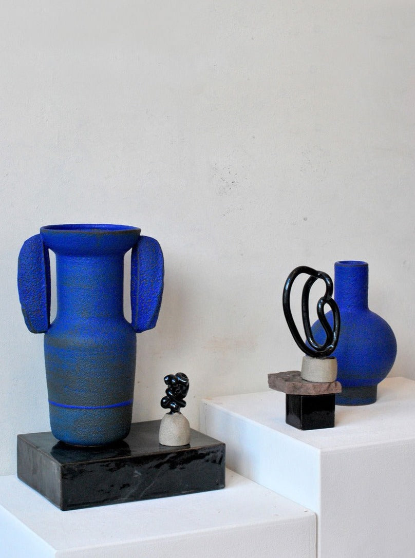 Two vibrant blue ceramic vases with unique designs on white pedestals, accompanied by smaller sculptures on blocks, against a plain white wall from the Moio Studio M702 collection.