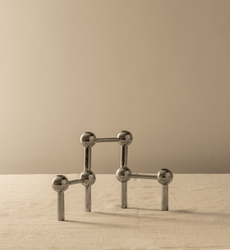 A minimalist sculpture featuring several interconnected silver spheres and rods forming a geometric structure on a pale surface against a light beige background, reminiscent of a Les Objets Amsterdam modular candle holder.