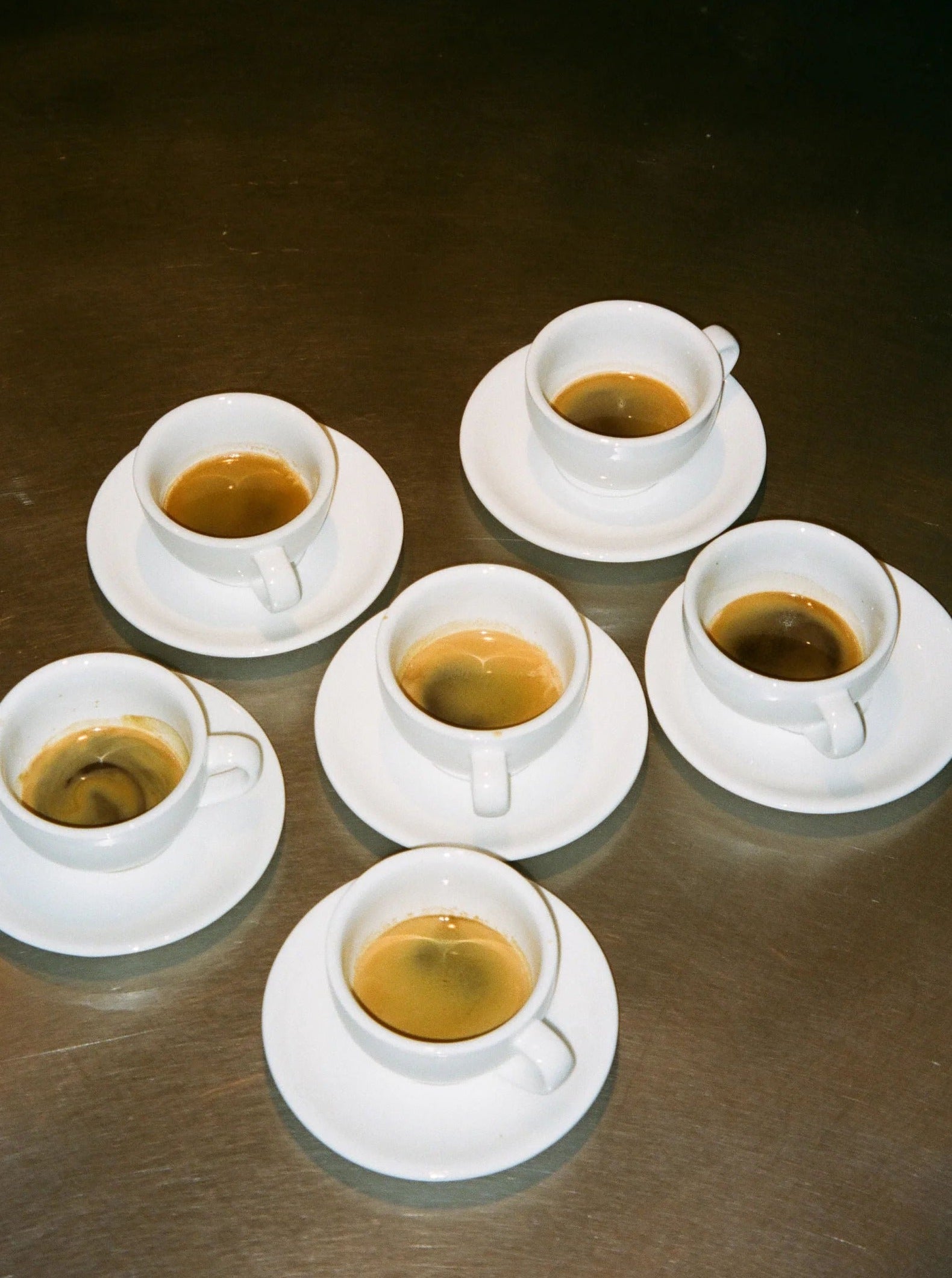 Five cups of espresso on saucers, arranged in a circle on a stainless steel table, each filled to varying levels with dark Service Projects Marlow coffee.