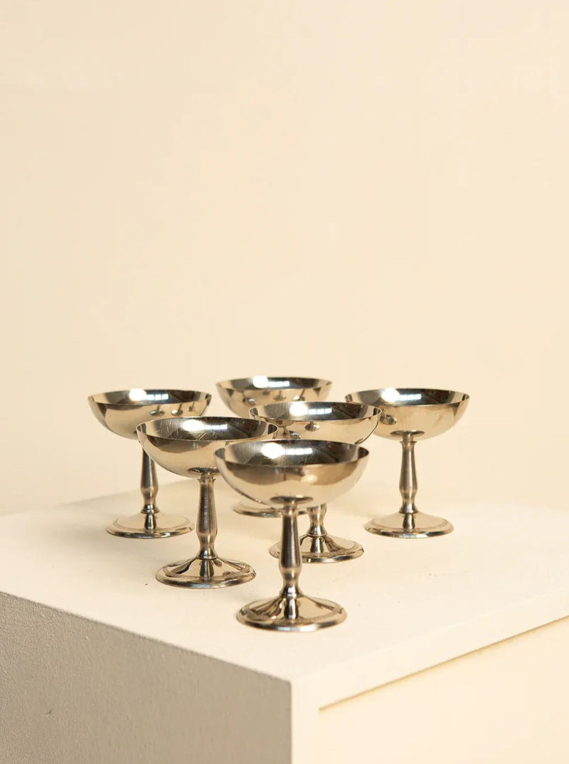 A set of six silver ice cream bowls and a 60's basket from Treaptyque are arranged on a light-colored pedestal against a beige background. The bowls have a classic stem and bowl design, reflecting light on their polished surfaces, exuding vintage charm reminiscent of 60s retro design.
