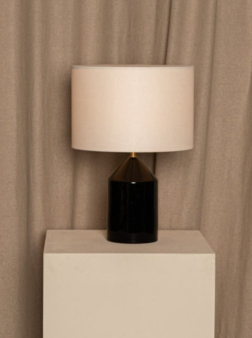 A stylish Simone & Marcel Josef Black Ceramic table lamp with a cotton drum lampshade stands on a neutral-colored pedestal against a draped beige curtain backdrop.