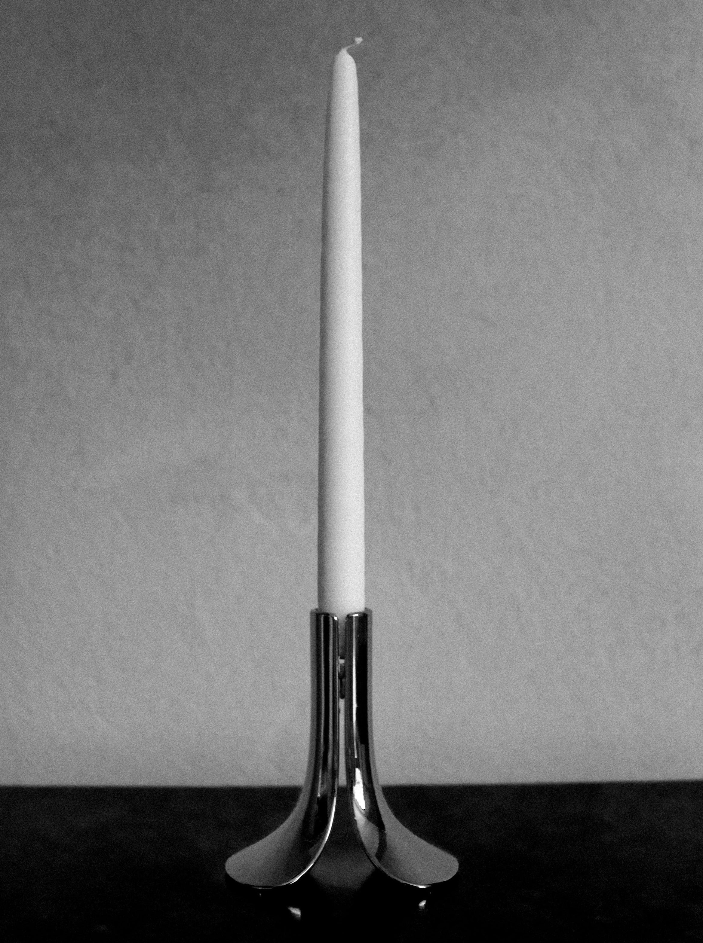 A black and white photo of an unlit white candle set in a sleek, Out For Lunch chrome candleholder placed on a tabletop against a plain background.