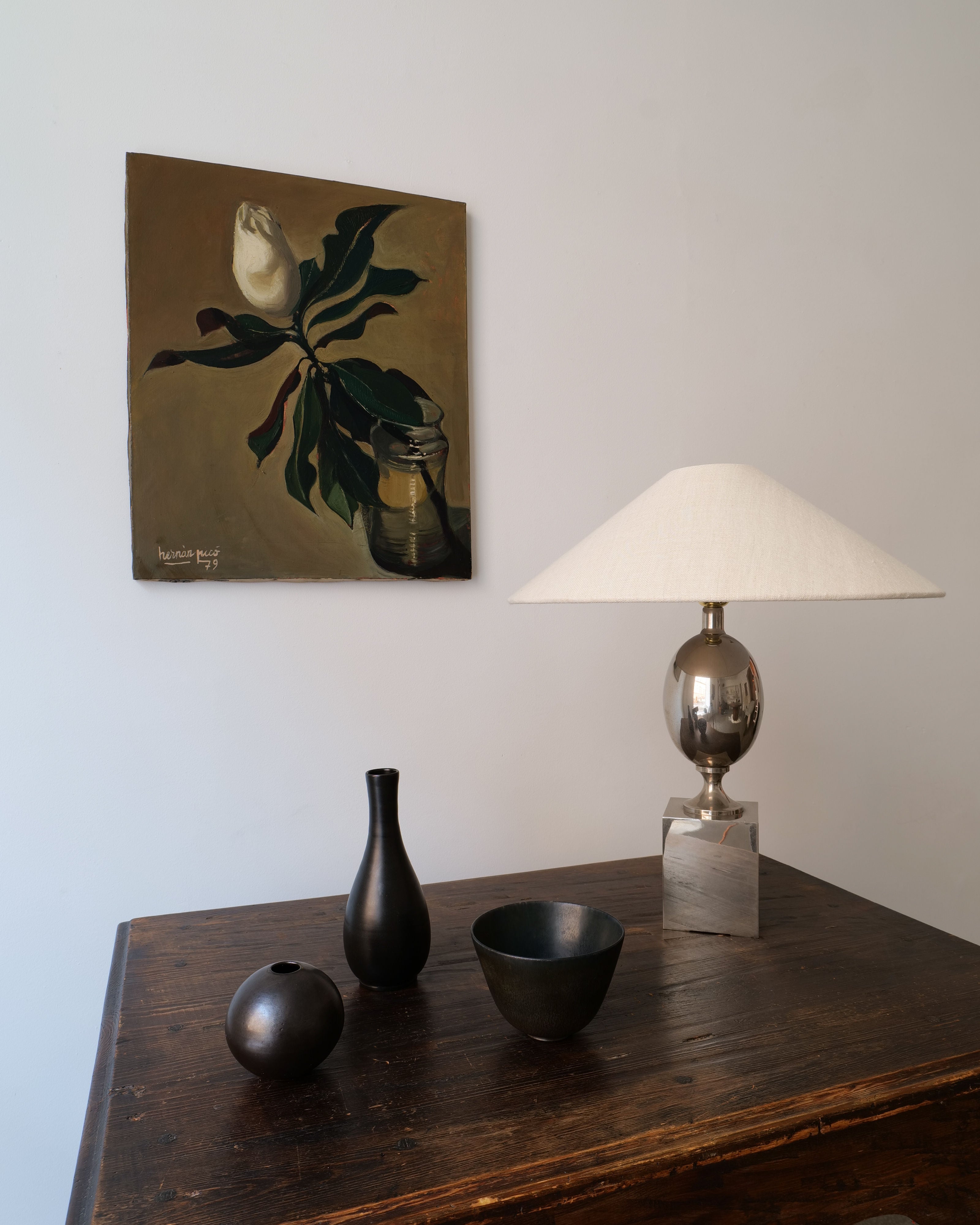 A rustic wooden table displaying a oil on canvas painting of a Magnolia Grandiflora 1979 stem in a jar, a silver table lamp with a white shade, and a black vase accompanied by a black bowl.
