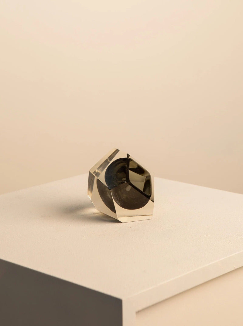 A transparent faceted Black "Diamant" Ashtray in Murano Glass by Flavio Poli for Seguso 60's displayed on a white pedestal against a neutral beige background. The ashtray casts a faint shadow on the surface. Created by Treaptyque.