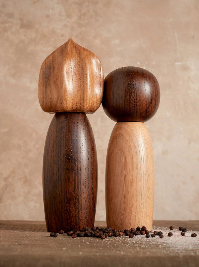 The LOBJET Picanto Salt + Pepper Mills stand side by side on a textured surface, showcasing contemporary design. The set includes a tall, dark mill with a round top and a shorter, lighter mill with a bulbous top, surrounded by scattered black peppercorns to enhance the luxury interior ambiance.