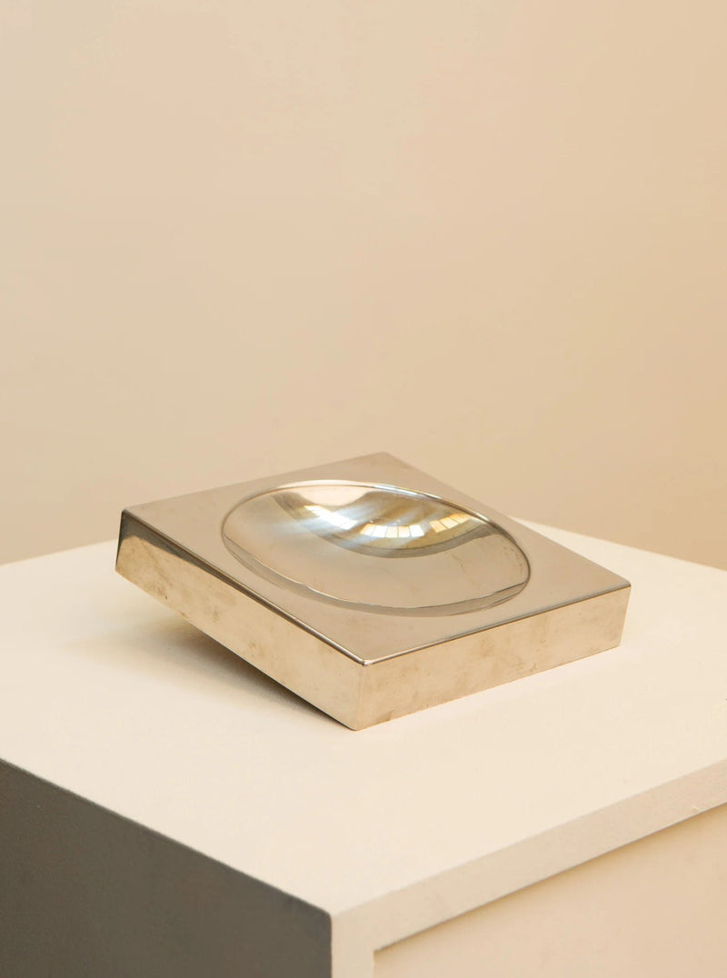 A sleek, silver Treaptyque 70's Square Metal Tray with a convex glass lens on top, displayed on a white pedestal against a neutral beige background.