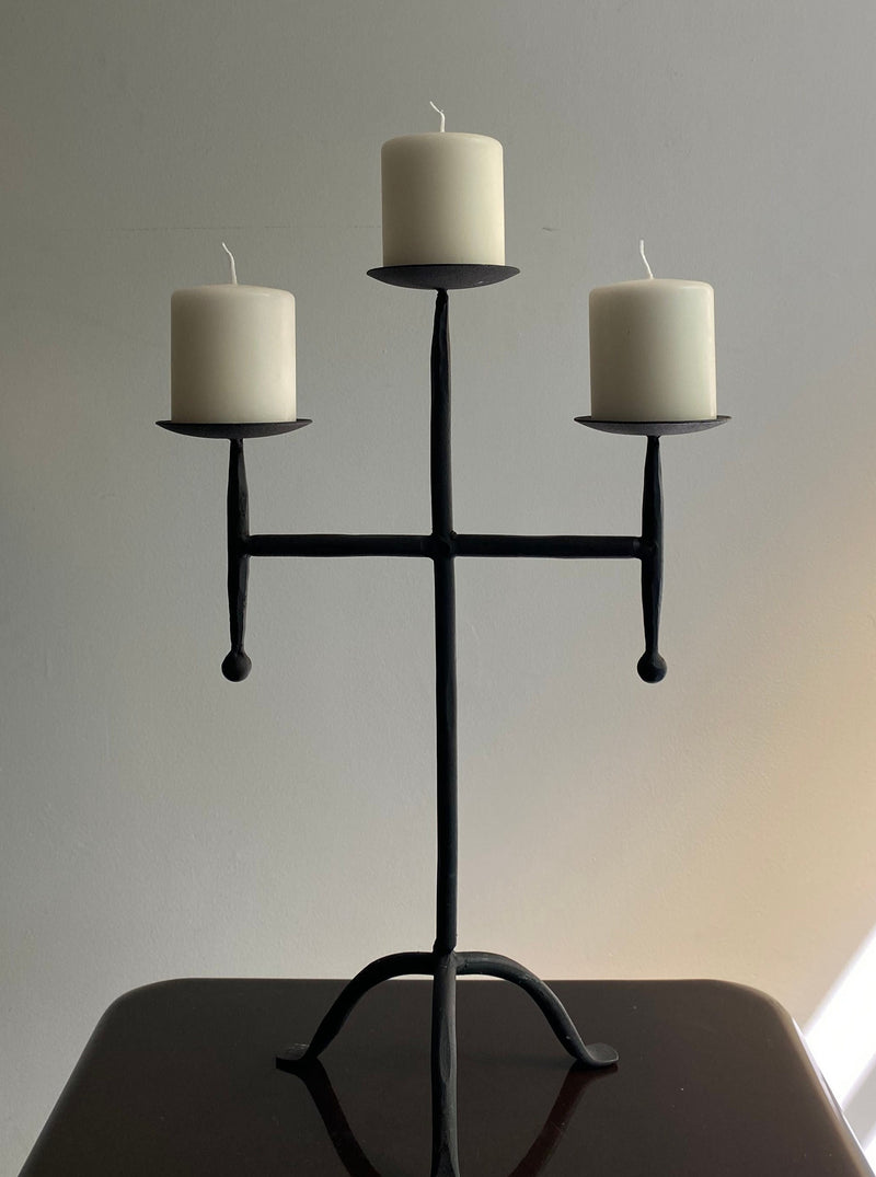 The Vintage Postmodern Iron Candle Holder by Les Objets Amsterdam is a sculptural black metal piece featuring three unlit white pillar candles. Crafted in the shape of a cross, it stands elegantly on a dark surface against a light wall, adding a luxurious touch to any interior.