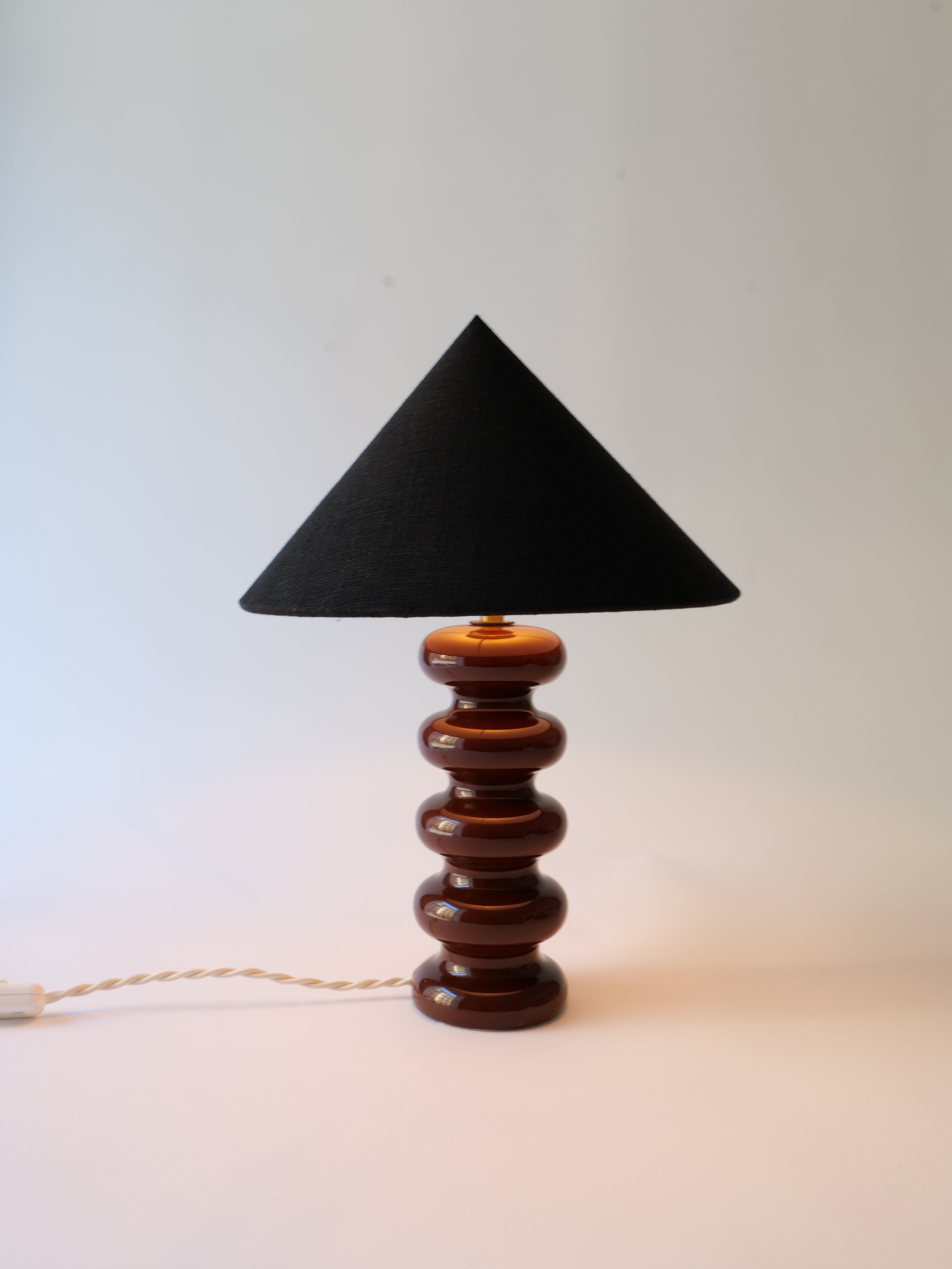 A modern Sculptural Ceramic Lamp from the Collection apart featuring a stack of glossy, brown, circular ceramic stones as its base, topped with a simple black, triangular lampshade. The lamp is connected to a white power.
