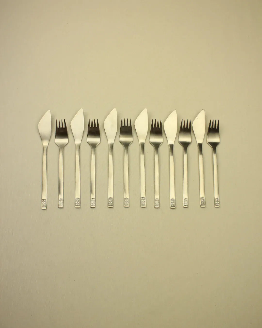 Nine Cutlery Set 70s Style forks with creatively designed cut-outs in their spades are arranged in a horizontal row against a plain beige background, featuring a minimalist design.