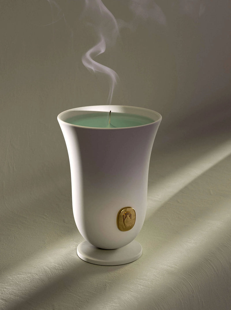 An elegant Bois Vert Candle by LOBJET in a white, tulip-shaped holder with a gold emblem, casting soft shadows on a textured beige surface. The candle's green wax is lit, emitting a thin stream of smoke.