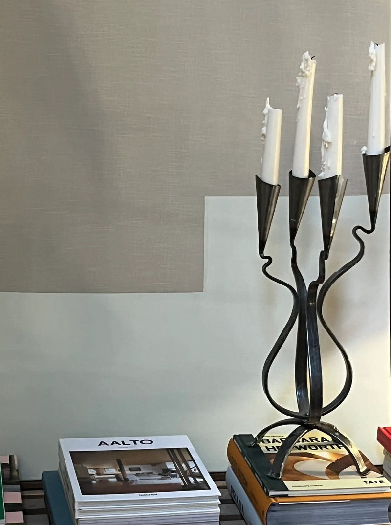 The Aesthete Label Post Modern Steel Large Candle Holder with five white candles sits on a stack of books beside a wall, while more books, including “AALTO,” are neatly arranged for striking post-modern decor.