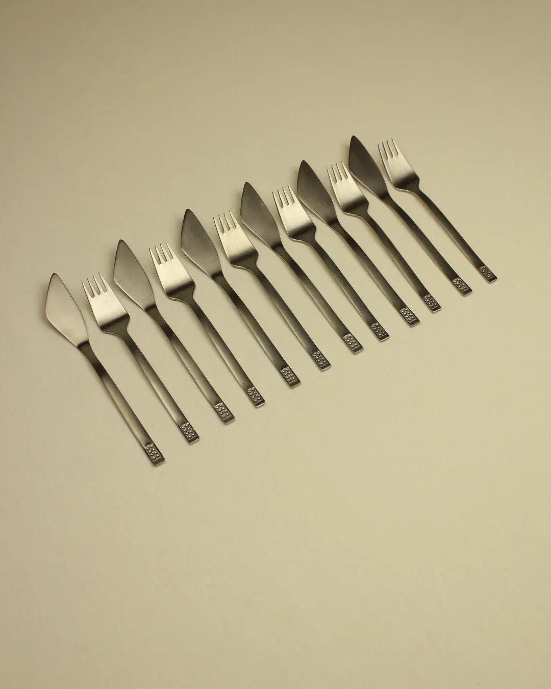 Eight satin stainless steel forks with unique square-ended handles and tined edges aligned diagonally across a cream background of the Cutlery Set 70s Style by Boga Avante Shop.