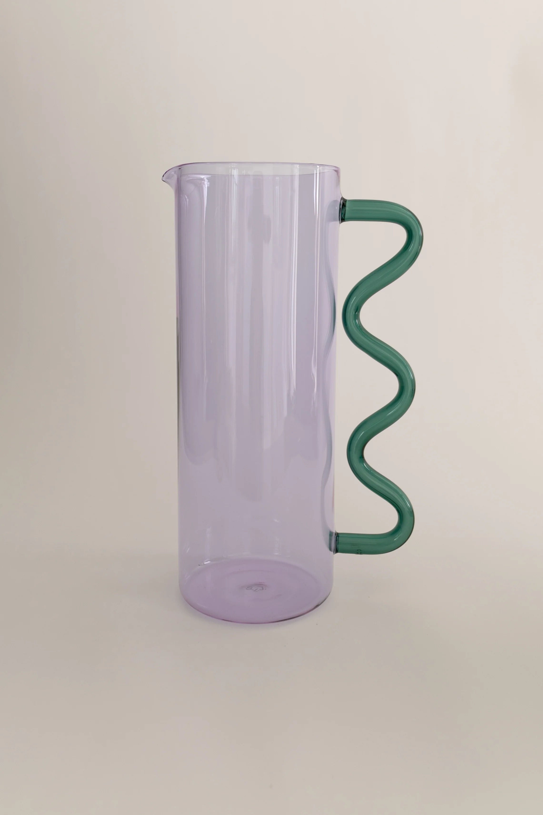 A lavender-tinted borosilicate glass Wave Pitcher Glass with a unique green, spiral handle standing against a light beige background by Sophie Lou Jacobsen.