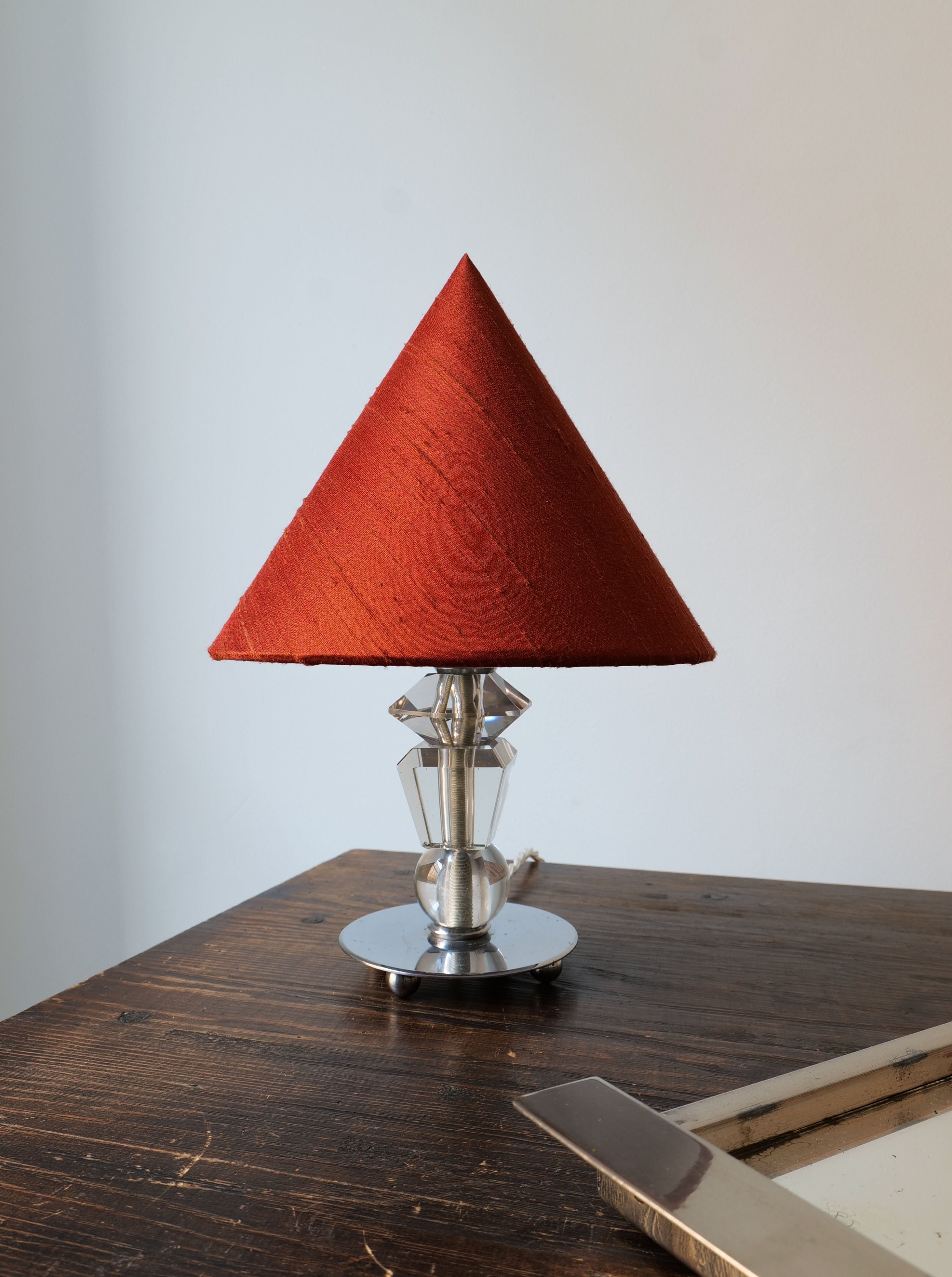 An Art Deco lamp with a red triangular lampshade on a chrome base, sitting on a wooden table against a white wall. The table also has a corner of a book visible.
An Collection apart Deco Crystal Table Lamp with collection apart's red triangular lampshade on a chrome base, sitting on a wooden table against a white wall. The table also has a corner of a book visible.