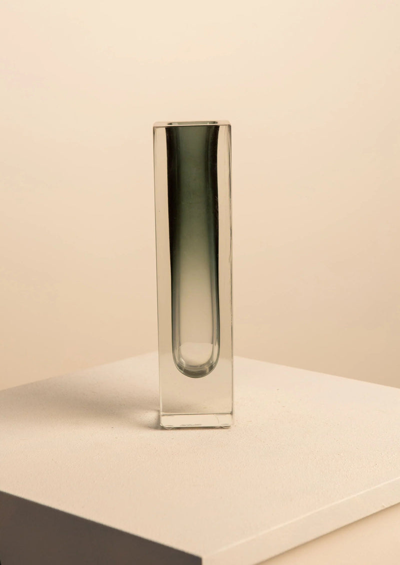The Black Vase by Flavio Poli for Seguso from Treaptyque, with a tall and narrow sculptural form and subtle dark gradient, stands out on a light surface against a plain beige backdrop, highlighting its minimalist aesthetic.